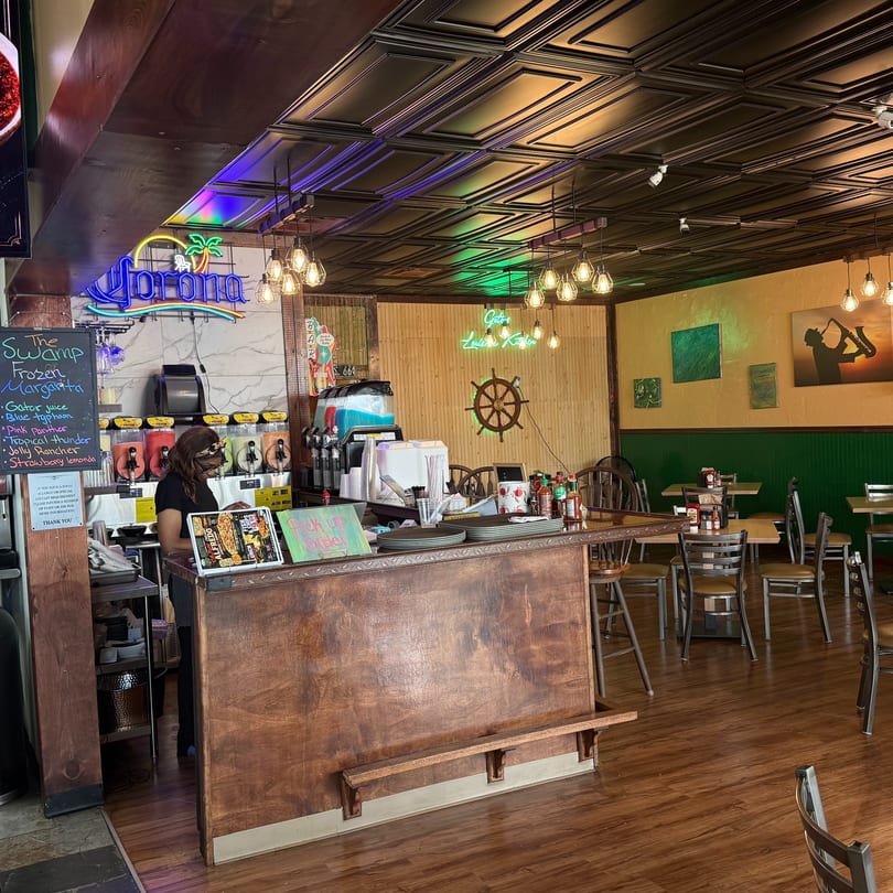 A restaurant interior with a wooden counter, margarita machines, and a woman standing behind the counter.